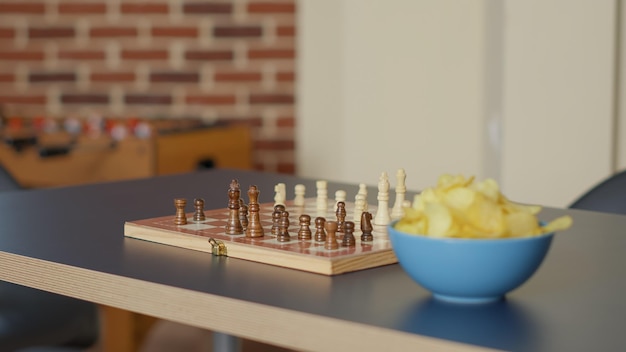 Mesa de sala vazia com jogo de tabuleiro de xadrez e tigela de batatas fritas, nenhuma pessoa no apartamento costumava se encontrar e se divertir com atividade estratégica. Ninguém no espaço com jogo de xeque-mate. Fechar-se.