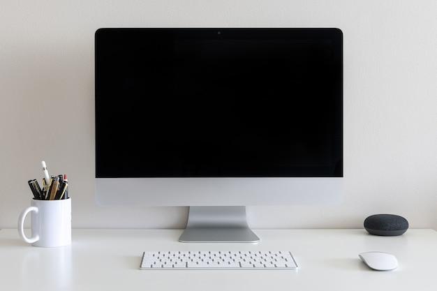 Mesa de trabalho com computador, copo com canetas e lápis contra uma parede branca