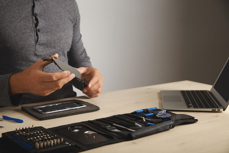 Foto grátis mestre segura uma nova tela para substituição acima do smartphone desmontado em seu laboratório, kit de ferramentas com instrumentos e laptop na frente dele na mesa branca, espaço para seu texto à direita