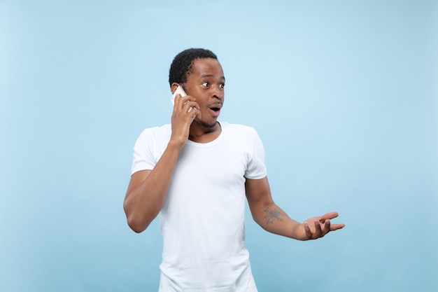 Metade do comprimento fechar o retrato de um jovem afro-americano de camisa branca sobre fundo azul. emoções humanas, expressão facial, conceito de anúncio. falando ao telefone, segurando um smartphone.