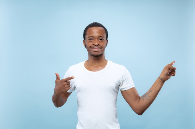 Metade do comprimento fechar o retrato de um jovem afro-americano de camisa branca sobre fundo azul. emoções humanas, expressão facial, conceito de anúncio. mostrando barra vazia, apontando, escolhendo, convidando.