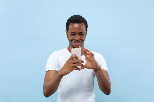 Metade do comprimento fechar o retrato de um jovem afro-americano de camisa branca sobre fundo azul. Emoções humanas, expressão facial, conceito de anúncio. Tirar uma foto ou conteúdo de vlog em seu smartphone.