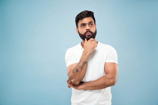 Metade do comprimento fechar o retrato de um jovem hindu de camisa branca sobre fundo azul. Emoções humanas, expressão facial, conceito de anúncio. Espaço negativo. Pensando enquanto segura a mão em sua barba. Escolhendo.