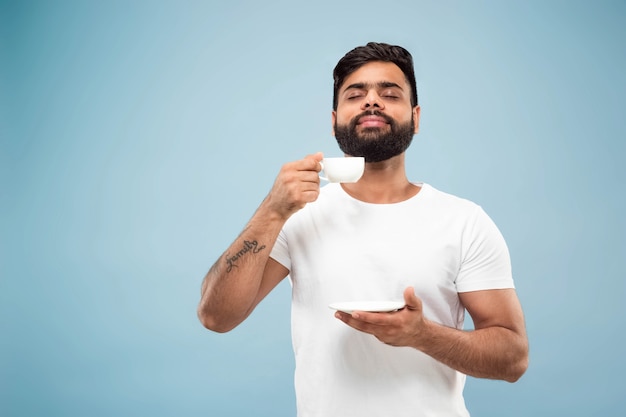 Metade do comprimento fechar o retrato de um jovem hindu de camisa branca sobre fundo azul. Emoções humanas, expressão facial, vendas, conceito de anúncio. Espaço negativo. Gostando de beber café ou chá.