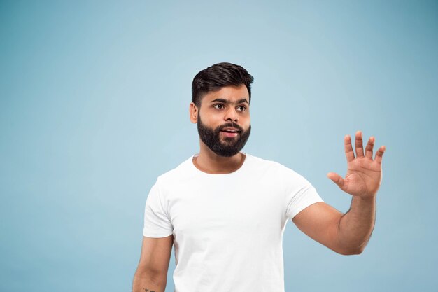Metade do comprimento fechar o retrato de um jovem hindu em uma camisa branca sobre fundo azul. Emoções humanas, expressão facial, conceito de anúncio. Espaço negativo. Mostrando a barra de espaço vazia, apontando, saudando.