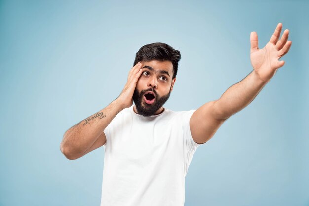 Metade do comprimento fechar o retrato de um jovem hindu em uma camisa branca sobre fundo azul. Emoções humanas, expressão facial, vendas, conceito de anúncio. Espaço negativo. Apontando para ficar chocado e surpreso.