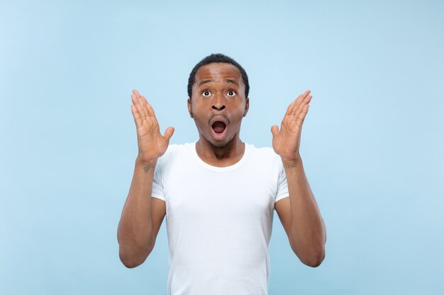 Metade do comprimento fechar o retrato do jovem modelo masculino afro-americano na camisa branca no espaço azul. Emoções humanas, expressão facial, anúncio ou conceito de aposta