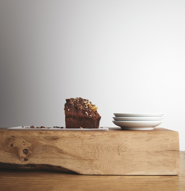 Metade do saboroso bolo de chocolate com frutas secas em um prato longo branco perto de três pequenos pratos de chá em branco em um tijolo cru de madeira e uma mesa grossa em um café