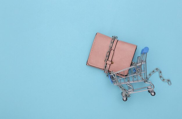 Mini carrinho de compras e carteira envolto em uma corrente de aço sobre fundo azul. vista do topo