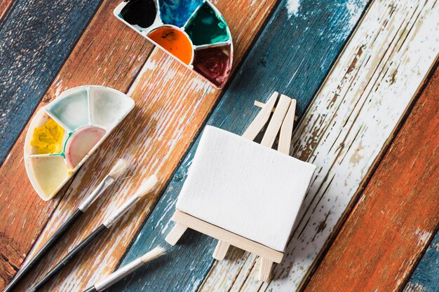 Mini cavalete em branco e equipamentos de pintura na velha mesa de madeira colorida