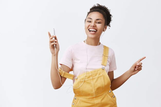 Mini concerto em meus ouvidos com novos fones de ouvido Brant. Retrato de uma mulher africana despreocupada e charmosa em um macacão estiloso, segurando um smartphone e dançando alegremente enquanto ouve música nos fones de ouvido