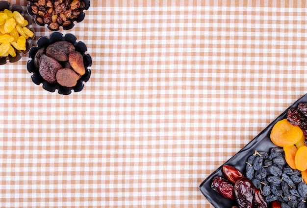 Mistura de datas secas de frutas passas damascos e cerejas em uma bandeja preta e em latas de mini torta na toalha de mesa xadrez com vista superior do espaço da cópia