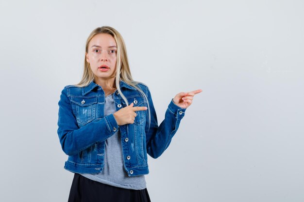 Moça apontando para o lado direito em camiseta, jaqueta jeans, saia e parecendo espantada.