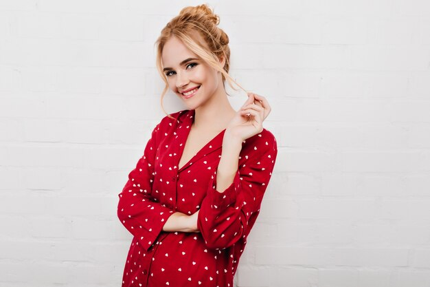 Moça bonita com maquiagem na moda, posando em pijamas vermelha e brincando com o cabelo loiro. Menina caucasiana sorridente de pijama em pé perto da parede de tijolos brancos.