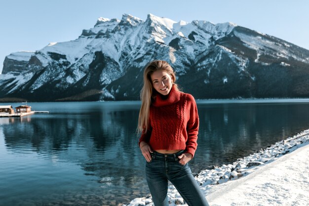 Moça bonita com sorriso branco em pé na praia perto do lago. Montanhas cobertas de neve. Vestindo blusa de malha vermelha e jeans azul. Cabelo longo loiro, sem maquiagem.