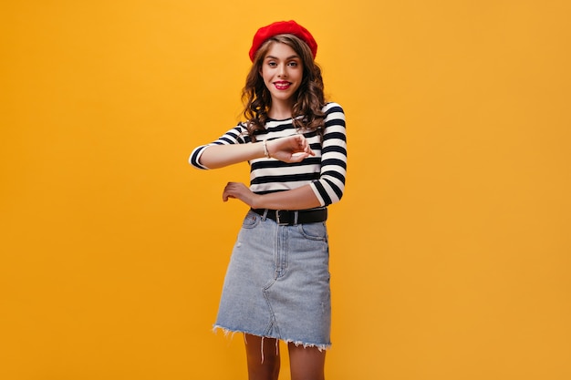 Moça bonita em poses de boina vermelha em fundo laranja. Jovem encantadora com cabelos ondulados em saia jeans, olhando para a câmera.