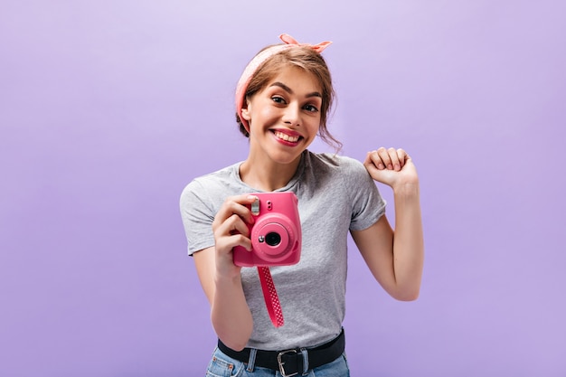 Moça bonita em poses de camiseta cinza com câmera rosa. mulher jovem e encantadora com roupas da moda e bandana legal, sorrindo sobre fundo isolado.