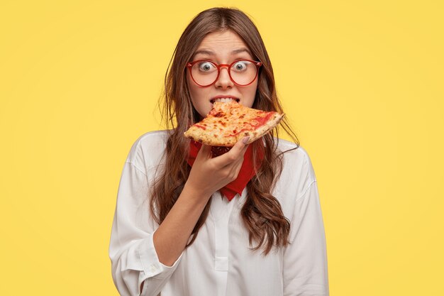 Moça bonita emotiva morde pizza deliciosa, olha direto na hora do lanche, visita pizzaria, surpreende-se com preços baixos, modelos sobre parede amarela. Pessoas, fast food e nutrição