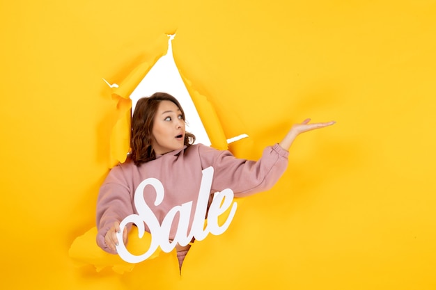 Moça bonita mostrando sinal de venda e apontando algo no lado esquerdo sobre fundo amarelo rasgado.