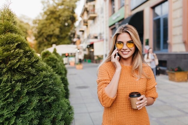 Moça bonita rindo falando ao telefone enquanto caminhava por arbustos com uma xícara de chá