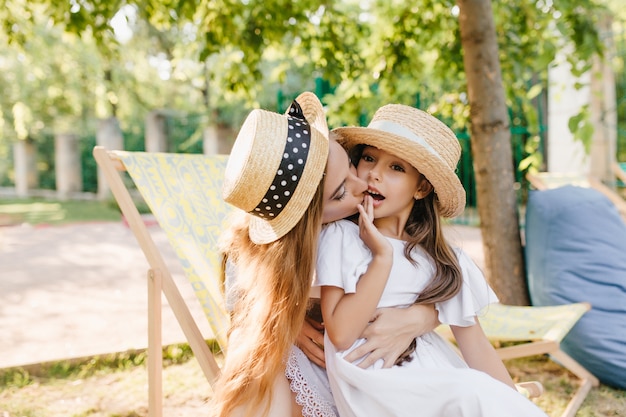 Moça bonita sentada na chaise-longue e segurando a filha nos joelhos, aproveitando um bom dia de verão. Retrato ao ar livre de uma linda mulher com chapéu vintage beijando a menina na bochecha.