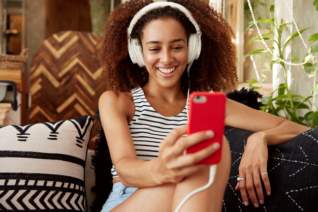 Moça bonita tem penteado afro, faz videochamada pelo smartphone e fones de ouvido, fala com um amigo online enquanto se senta no confortável sofá com almofadas.