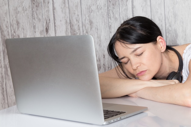 moça de camisa branca com earphoens pretos adormeceu na frente do laptop em cinza