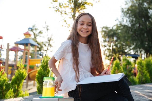 Moça de sorriso que senta-se ao ar livre com caderno e suco