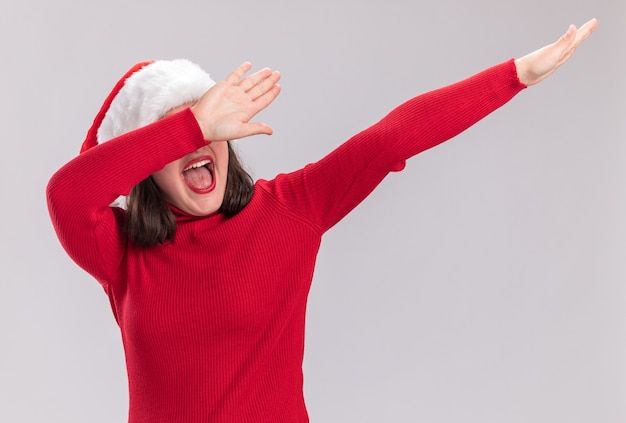 Moça engraçada de suéter vermelho e chapéu de Papai Noel gesticulando com as mãos felizes e animadas em pé sobre um fundo branco
