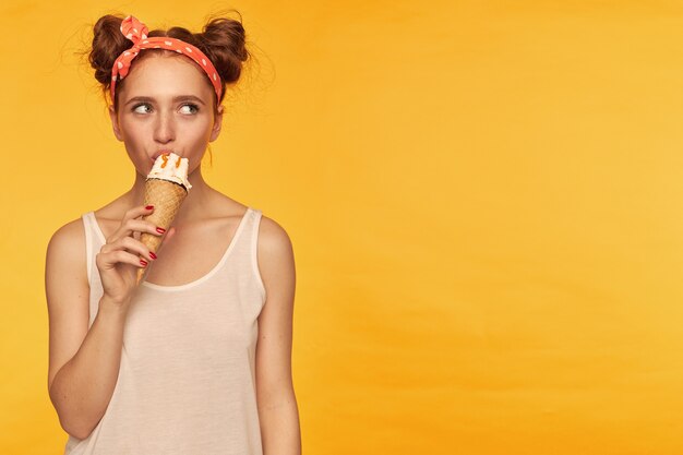 Foto grátis moça, linda mulher ruiva com dois pães. vestia um top branco e uma faixa vermelha pontilhada no cabelo tomando um sorvete. observando à direita no espaço da cópia, isolado sobre a parede amarela