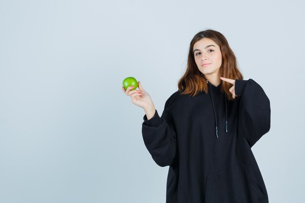 Moça segurando maçã enquanto aponta para o lado esquerdo em um capuz grande, calças e está bonita. vista frontal.
