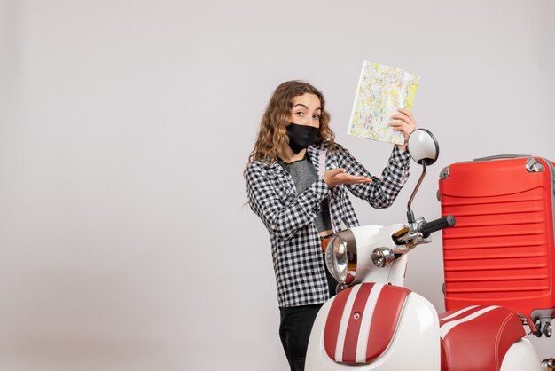 moça simpática com máscara segurando mapa em pé perto de motocicleta com mala vermelha