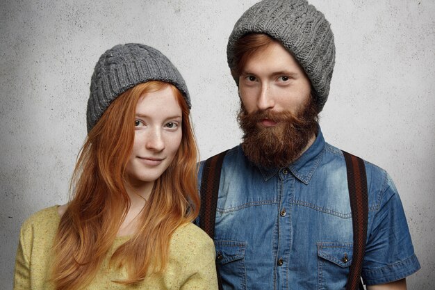 Moda de inverno. jovem casal caucasiano feliz usando chapéus de malha cinza quentes posando dentro de casa contra a parede.