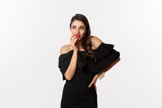 Moda e beleza. Mulher glamour em um vestido preto pensando, tendo uma ideia, em pé sobre um fundo branco.