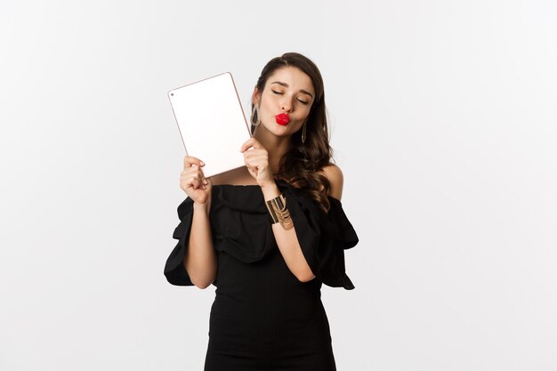 Moda e conceito de compras. Mulher linda com lábios vermelhos, vestida de preto, mostrando tablet digital e fazendo carinha de beijo, fundo branco