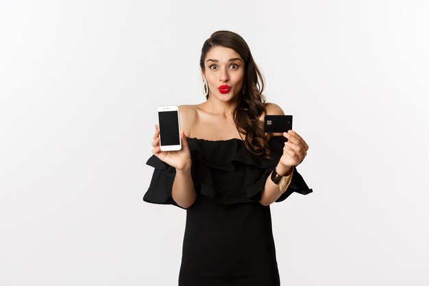 Moda e conceito de compras online. Mulher jovem feliz em um vestido preto, mostrando o cartão de crédito e a tela do celular, em pé sobre um fundo branco.