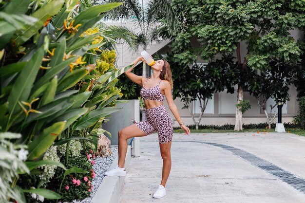 Moda elegante e jovem caucasiana em forma de mulher desportiva com top de cami de leopardo e shorts de motociclista do lado de fora segurando um shaker de proteína e uma garrafa de água na mão