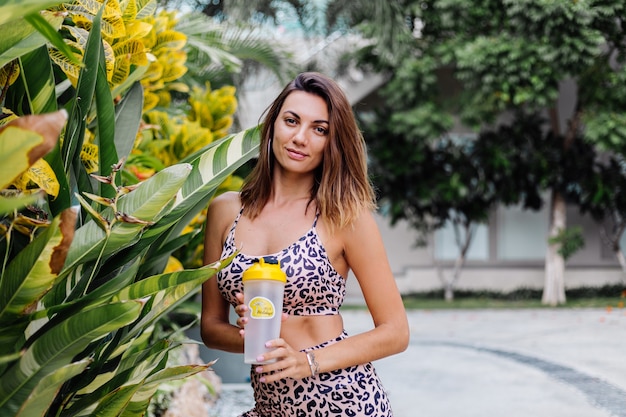 Moda elegante e jovem caucasiana em forma esportiva com blusa de cami leopardo e shorts de motociclista do lado de fora segurando um shaker de proteína