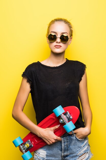 Moda garota muito legal em óculos de sol e skate sobre fundo colorido amarelo