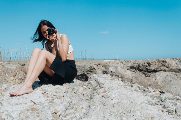 Moda jovem mulher tirando foto com a câmera sentada na rocha ao ar livre