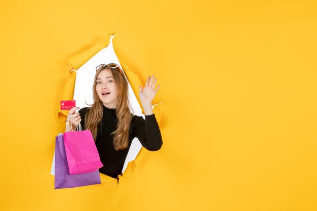 Moda jovem segurando sacolas de compras e cartão de crédito pelo buraco de papel rasgado na parede