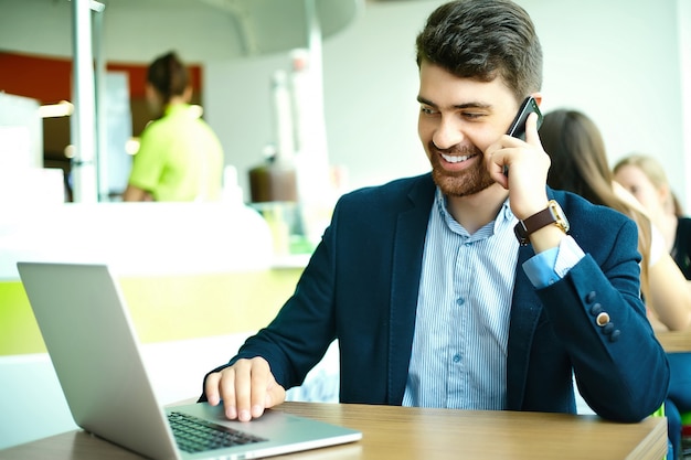 Moda jovem sorridente homem hipster no café da cidade durante a hora do almoço com o notebook no terno falando no telefone