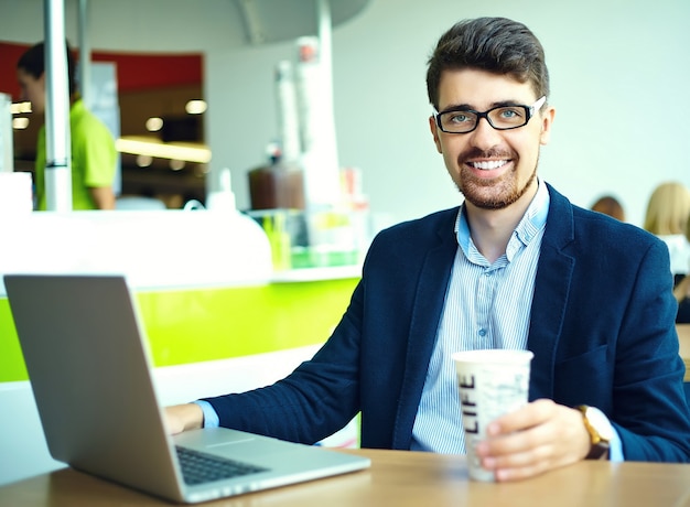 Moda jovem sorridente homem hipster tomando café no café da cidade durante a hora do almoço com o notebook no terno