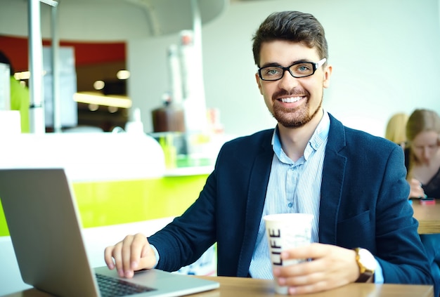 Moda jovem sorridente homem hipster tomando café no café da cidade durante a hora do almoço com o notebook no terno