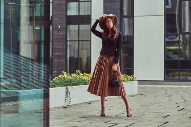 Moda mulher elegante vestindo uma jaqueta preta, chapéu marrom e saia com uma embreagem de bolsa andando no centro da cidade europeia.