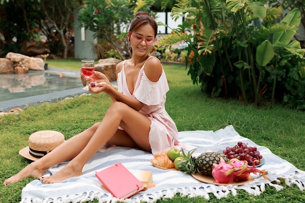 Modelo asiático bastante gracioso sentado no cobertor, bebendo vinho e desfrutando de piquenique de verão no jardim tropical.