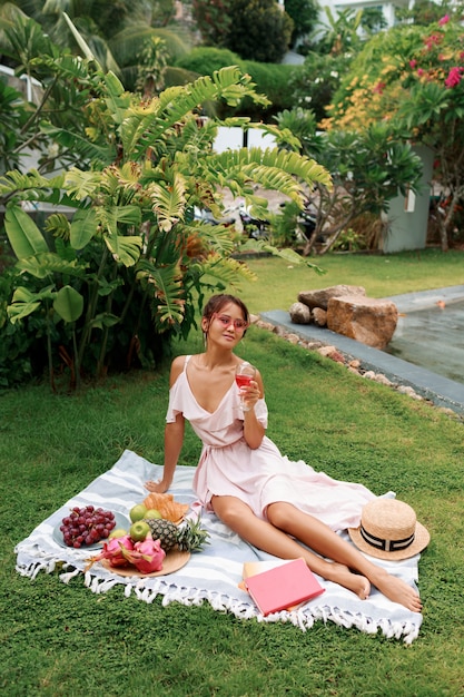 Modelo asiático gracioso romântico sentado no cobertor, bebendo vinho e desfrutando de piquenique de verão no jardim tropical.