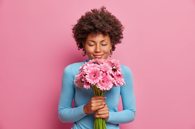 Modelo atraente de pele escura e satisfeita recebe flores como presente, fica com os olhos fechados, desfruta de suas gerberas favoritas,