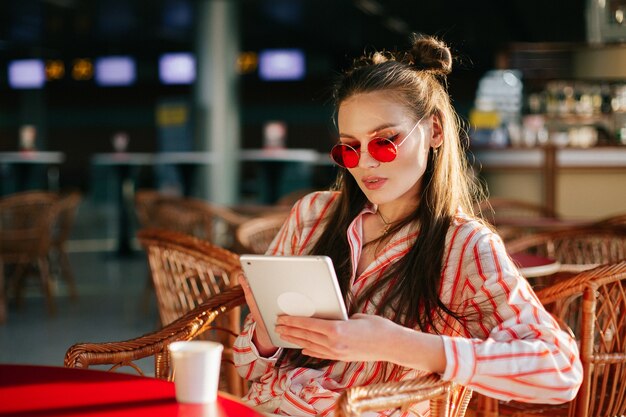 Modelo bonito em óculos de sol vermelhos trabalha com seu tablet sentado no café