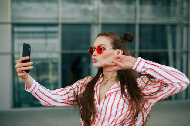 Modelo bonito em óculos vermelhos leva selfie em seu smartphone